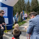 Grupa ludzi przy niebieskim balonie na świeżym powietrzu. Mężczyzna wręcza medale dzieciom. W tle zalesione otoczenie.
