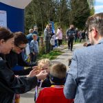 Grupa osób stoi na ścieżce, dzieci otrzymują medale. W tle zielone drzewa i niebieskie plakaty. Dzień jest słoneczny.