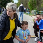 Na zdjęciu starsza kobieta i dwaj chłopcy z medalami na szyi. Kobieta ma żółtą koszulkę, dzieci są ubrane na niebiesko. W tle widać ludzi i drzewa, chodnik i niebieską poręcz.