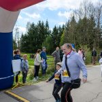 Kobieta z chłopcem biegnie podczas zawodów, mijając niebieski balon startowy. W tle grupa osób przy drodze. Otoczenie jest zielone z drzewami i trawą.
