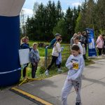 Na zdjęciu widać dzieci w sportowych strojach na zewnątrz. W tle są drzewa i kilka osób, obok niebieskiej konstrukcji. Pogoda jest słoneczna.