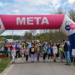 Grupa dzieci i dorosłych stoi na linii startu biegu pod różowo-niebieskim balonem z napisem META na tle drzew.