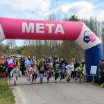 Dzieci biegają spod różowego nadmuchanego łuku mety. W tle drzewa i widzowie. Niebieska i biała kolorystyka dekoracji.