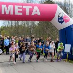 Na zdjęciu dzieci startują w biegu pod różowym łukiem z napisem META na tle lasu. Tłum kibiców w kolorowych ubraniach obserwuje wydarzenie. Dzień jest słoneczny, a niebo pochmurne.