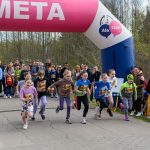 Grupa dzieci w kolorowych strojach biegnie przez linię mety. Tło stanowi las i tłum kibiców. Nad metą różowy łuk z napisem META.
