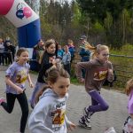 Grupa dzieci biegnie w wyścigu na tle lasu. Nosi kolorowe koszulki i numery startowe. Za nimi stoi tłum kibiców i duży kolorowy balon.