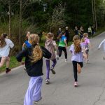 Grupa dzieci biegnie po asfaltowej drodze przez las. Mają kolorowe ubrania, a wokół jest zieleń drzew. Tło wypełnia leśna sceneria z obserwującymi ludźmi.