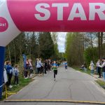 Biegaczka mija różowy łuk z napisem START. Po bokach stoją drzewa i kibicujący ludzie. Pogoda jest słoneczna.