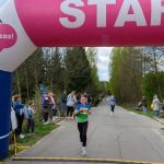 Biegaczki przebiegają pod różowym łukiem startowym na asfaltowej drodze. Wokół widoczni kibice i zielone drzewa w tle. Pogoda jest pochmurna, atmosfera sportowa.