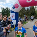 Zawody biegowe na wiejskiej drodze, dzieci w niebieskich koszulkach na pierwszym planie. Tło to drzewa i kibice. Nad nimi kolorowy łuk z napisem Ale Czas!.