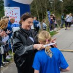 Na zdjęciu kobieta zakłada medal dziecku w niebieskiej koszulce. W tle ludzie kibicują wydarzeniu na świeżym powietrzu. Dookoła drzewa, asfaltowa droga i plakaty promocyjne.