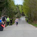 Na zdjęciu grupa dzieci biegnie po asfaltowej drodze otoczonej drzewami. Po lewej stronie stoi kilka osób, niebo jest jasne. Las w tle jest zielony.