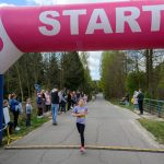Na zdjęciu widzimy młodą biegaczkę przekraczającą linię startu. Nad drogą znajduje się różowo-biały łuk z napisem START. W tle widać ludzi obserwujących wydarzenie i zielone drzewa.