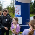 Grupa osób stoi na zewnątrz przy niebieskim filarze z mapą. Tło to zielone drzewa i wiejskie zabudowania. Dzieci rozmawiają z dorosłymi.