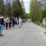 Na zdjęciu widoczny jest bieg uliczny dzieci i kibice. Przy drodze rosną zielone drzewa. Dzień jest jasny, a niebo lekko zachmurzone.