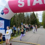 Na zdjęciu dzieci biegną przez bramę startową podczas wyścigu. Kolory dominujące to różowy i biały na bramie. W tle widoczni są kibice i zalesiona okolica.