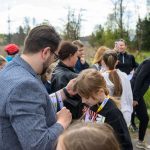 Grupa ludzi, w tym dzieci na zewnątrz. Mężczyzna w marynarce przypina coś dziewczynce. W tle drzewa i plakat na niebieskiej kolumnie.