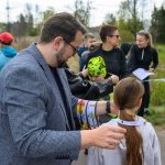 Grupa osób na świeżym powietrzu, mężczyzna w marynarce zakłada identyfikator na dziewczynkę. Tło to zieleń i drzewa. Osoby noszą sportowe ubrania.