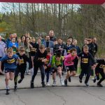 Grupa młodych biegaczy startuje w wyścigu, ubrani w kolorowe stroje. W tle widać kibiców. Otoczenie jest zielone z drzewami.