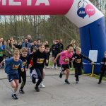 Dzieci startują w biegu pod różowo-niebieskim łukiem z napisem META. Tło stanowi zielony las. Obok stoi mężczyzna z mikrofonem.