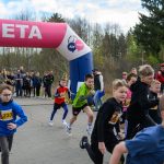 Młodzież biegnie przez różowy łuk z napisem META. W tle las i grupa kibiców. Dzieci mają różnokolorowe stroje sportowe.