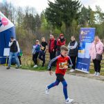 Chłopiec biegnie w zawodach na tle lasu. Ludzie stoją obok niebiesko-różowego łuku i plakatu. Chłopiec ma czerwoną bluzę i niebieskie legginsy.