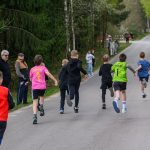 Grupa dzieci w kolorowych strojach biegnie po asfaltowej drodze otoczonej zielonym lasem. W tle widać ludzi stojących przy drodze. Dzień jest pochmurny, ale jasny.