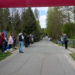 Droga w lesie otoczona kibicami; biegacze na trasie. Wiosna, dominują zielenie i błękity. Nad drogą łuk startowy.