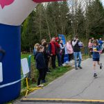 Chłopiec biegnie ulicą w zawodach, a widzowie kibicują przy drodze. Nad nim znajduje się kolorowy, dmuchany łuk. Tło tworzy zielony las.