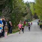 Dzieci biegną w wyścigu ulicznym. Tło stanowi zielony las. Po lewej stronie stoi grupa widzów.