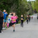 Uczestnicy biegu dziecięcego na asfaltowej drodze otoczonej drzewami. Przód zajmuje chłopiec w różowej koszulce, nr 235. W tle kibicują rodziny.