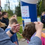 Mężczyzna zakłada medal na szyję chłopca w różowej bluzie. W tle widoczni są ludzie i zielone drzewa. Scena jest na świeżym powietrzu.