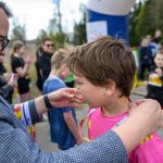 Mężczyzna w okularach zakłada medal chłopcu w różowej koszulce. W tle inne dzieci i dorośli. Sceneria zewnętrzna z drzewami.