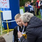 Chłopiec w czarnej bluzie na medal otrzymuje nagrodę od kobiety w kurtce. W tle widoczni są uczestnicy zawodów. Scena rozgrywa się na zewnątrz, obok dużego niebieskiego balonu.