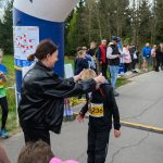 Na zdjęciu jest bieg młodzieżowy na zewnątrz. Kobieta zakłada medal chłopcu w czarnym stroju z numerem 236. W tle są drzewa i kibice.
