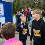 Na zdjęciu widoczna jest grupa dzieci w ciemnych strojach z numerami 233 i 234. W tle widać kibicujących ludzi oraz drzewa. Młoda kobieta w czarnej kurtce zapisuje wyniki na dużym niebieskim stojaku.