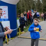 Chłopiec w niebieskiej bluzie biegnie w wyścigu. W tle grupa ludzi stoi przy drodze, niektórzy z medalami. Po lewej stronie widoczny jest duży niebieski balon reklamowy.