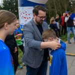 Mężczyzna zakłada medal chłopcu w niebieskiej bluzie. W tle grupa ludzi na dworze, drzewa i plansza z mapą. Pogoda jest pochmurna.