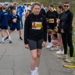 Młoda dziewczyna w czarnym stroju stoi z numerem startowym na tle uczestników biegu. W tle widoczne drzewa i samochody. Inni biegacze przygotowują się, wiążąc buty na asfalcie.