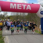 Biegacze startują spod różowo-białej bramy z napisem META. W tle drzewa i widownia. Pogoda jest pochmurna.