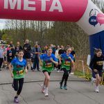 Na zdjęciu grupa młodych biegaczy startuje spod różowo-białego łuku z napisem META. Tło stanowi zielony las i zebrani kibice. Atmosfera zawodów sportowych na ścieżce asfaltowej.