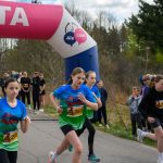 Dzieci startują w wyścigu spod różowego łuku z napisem 'META'. Mają kolorowe koszulki, a w tle widać drzewa i widzów. Atmosfera biegu i emocji na świeżym powietrzu.