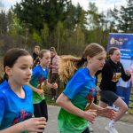 Kilka dziewcząt biegnie w wyścigu, ubrane w niebieskie koszulki. W tle widoczne są drzewa i kibice. Obok stoi niebieski baner reklamowy.