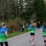 Cztery osoby biegną wzdłuż asfaltowej drogi w lesie; mają kolorowe koszulki. Tło to zieleń drzew i krzewów. Kilka osób obserwuje bieg.