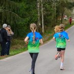 Cztery osoby biegną po asfaltowej drodze, ubrane w kolorowe niebiesko-zielone koszulki. W tle widać drzewa i parę osób obserwujących wydarzenie. Atmosfera jest sportowa i pełna energii.