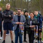 Grupa ludzi stoi na tle drzew, trzymając medale. Są ubrani kolorowo. Tło jest zielone, jak w parku na wiosnę.