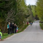 Biegacze na asfaltowej drodze w otoczeniu zielonego lasu, obserwowani przez grupę osób. Po bokach drogi widoczne drzewa. Chmury na niebie.