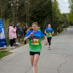 Na zdjęciu widać biegacza w niebiesko-zielonej koszulce. W tle jest widoczna droga i drzewa. Kilka osób kibicuje przy trasie.