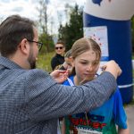 Mężczyzna zakłada nastolatce medal na niebieskiej koszulce. Tło to wiejska droga z drzewami i niebiesko-białym dmuchanym łukiem. W oddali widać grupkę osób.