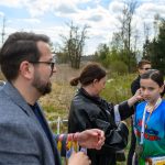 Na zdjęciu widoczne jest dziecko w niebieskiej koszulce z medalem. W tle dorosłe osoby i zielone drzewa. Scena ma miejsce na świeżym powietrzu, z widocznymi chmurami na niebie.